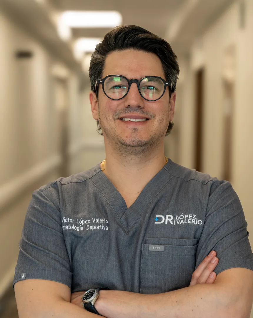 Retrato del Dr. Víctor López Valerio, traumatólogo y ortopedista, con bata quirúrgica gris y los brazos cruzados